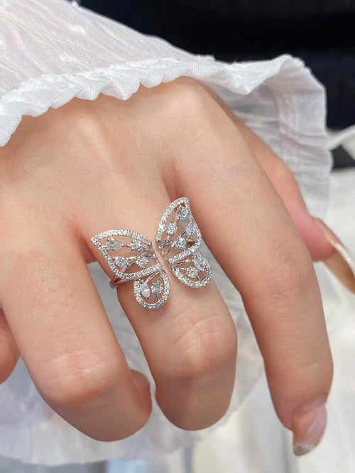 A silver butterfly ring set with clear crystals, worn on a finger. The hand is adorned with a white lace sleeve, and the background is softly blurred.