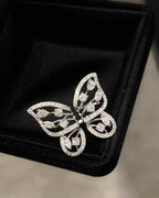 A silver butterfly-shaped brooch adorned with pavé-set clear stones, displayed on a black velvet jewelry box.