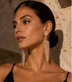 A woman with dark hair in a low bun wears large feather-shaped drop earrings with a textured pavé setting. She is posed against a warm, textured stone wall under dramatic, directional lighting.