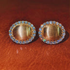 A pair of gold oval stud earrings with a halo of small blue iridescent crystals, displayed on a textured brown leather surface.