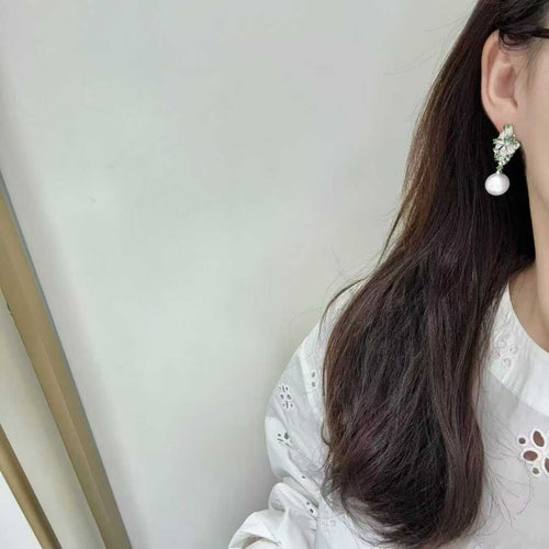 A close-up side view of a woman wearing a dangling earring with a white pearl and a silver-toned crystal-embellished top, paired with a white eyelet blouse.