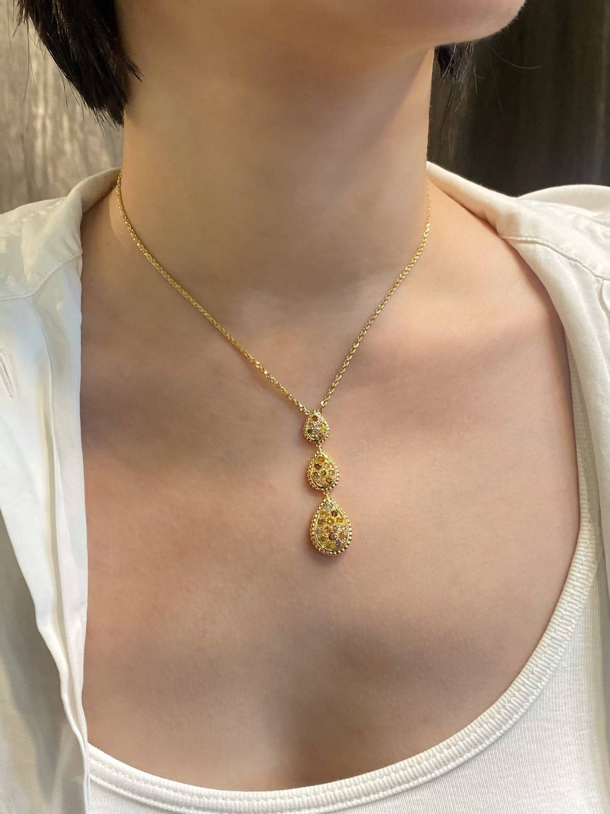 A gold pendant necklace with three stacked teardrop-shaped charms, worn on the neck of a person wearing a white top.