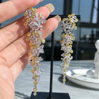 A close-up of a hand holding a pair of long, ornate drop earrings with silver-toned and yellow-gold floral clusters, displayed against a softly blurred indoor background with a black jewelry stand.