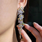 A close-up of a person wearing an ornate, long drop earring adorned with yellow and clear stones in a silver-toned setting, with fingers gently touching the lower part of the earring.