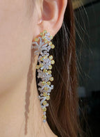 A close-up of a woman's ear wearing a dramatic, long dangle earring. The earring is made of silver-toned metal and yellow gold-toned metal, heavily set with clear gemstones and accented with small round yellow gemstones.