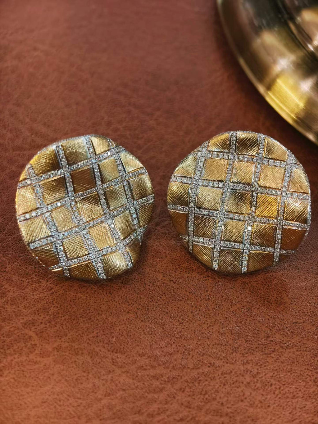 A pair of round gold-toned earrings with a woven grid pattern outlined in silver-toned metal, displayed on a brown textured surface.